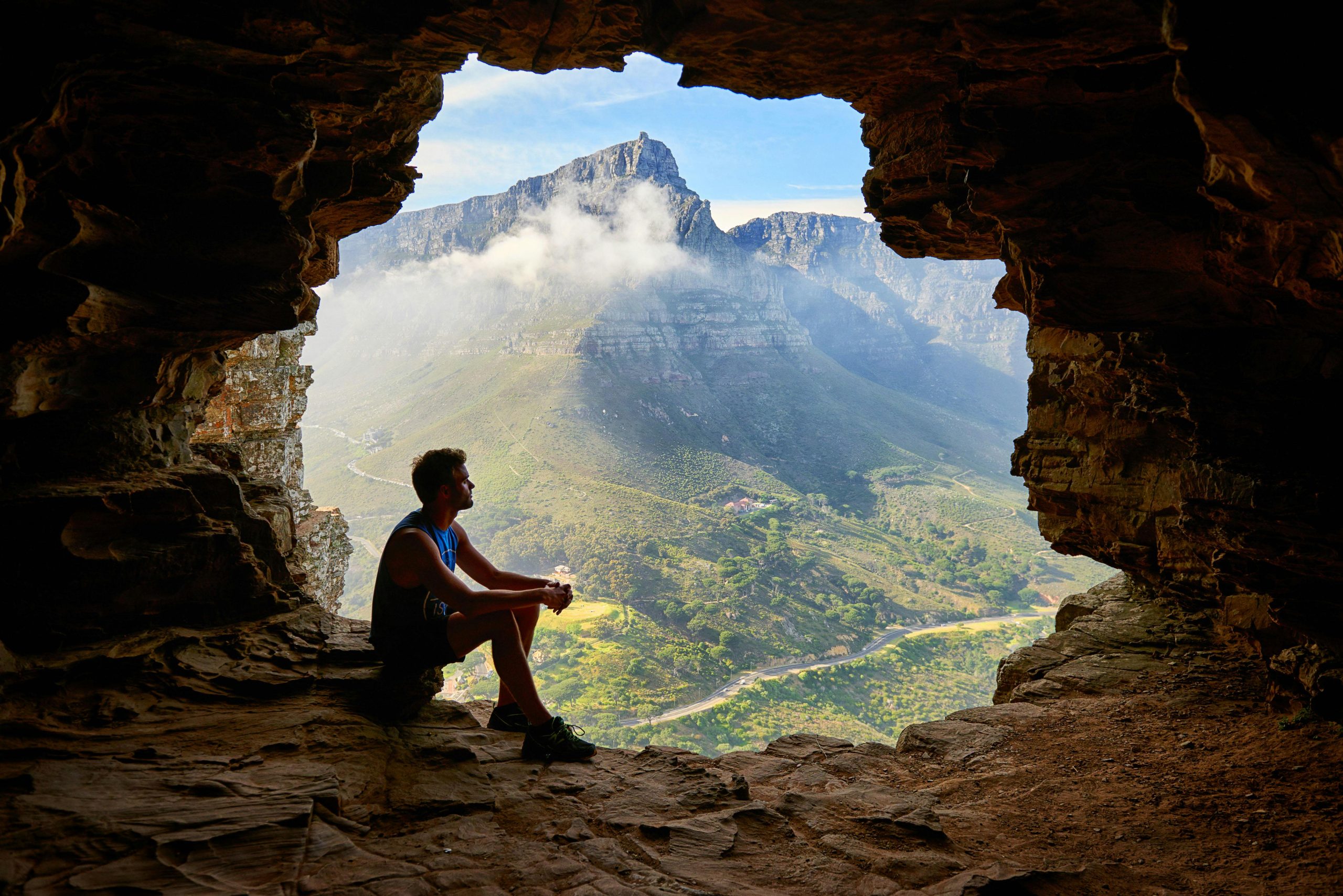 A man sitting inside a cave, gazing at a stunning mountain view with clouds surrounding the peaks